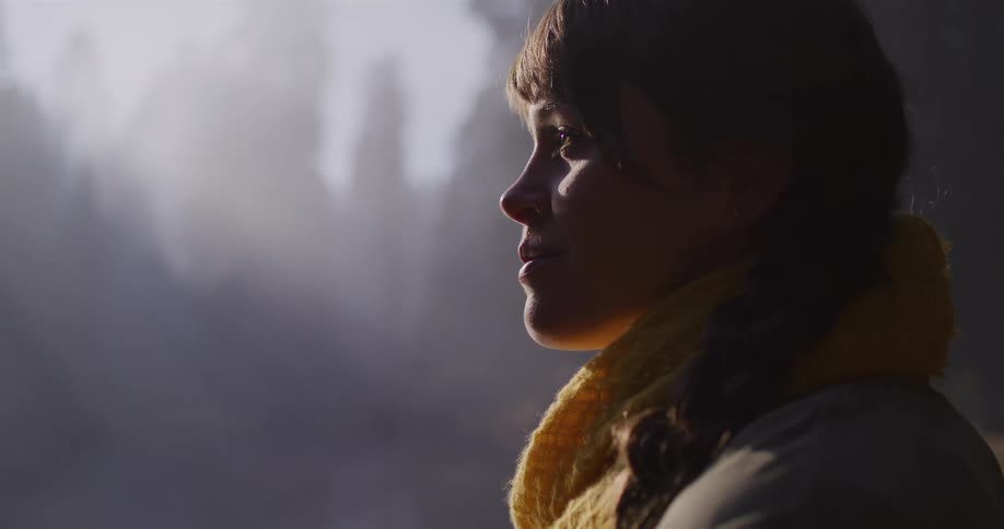 A young woman's profile is beautifully illuminated by sunlight as she gazes in wonder at a misty forest landscape in slow motion