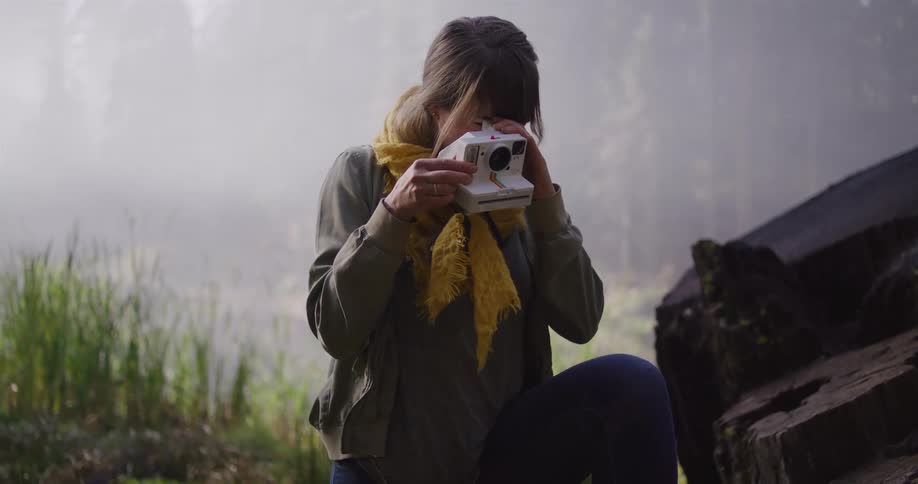 A young woman in a yellow scarf explores a misty forest, climbing over large rocks and stopping to take a picture with her instant camera