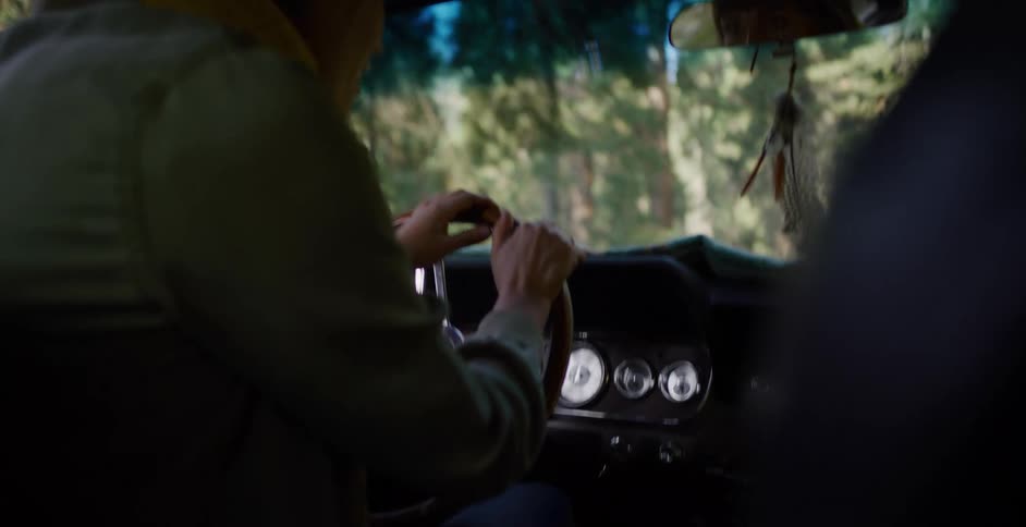 From the backseat, a woman drives a classic car with a wooden steering wheel down a bumpy road through a sunny forest