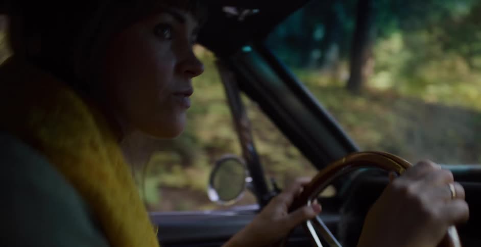 A woman wearing a yellow scarf enjoys a scenic drive through a sunlit forest in her classic car