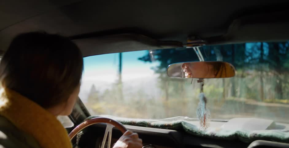 Over-the-shoulder shot of a woman smiling as she drives her classic car through a sun-dappled forest
