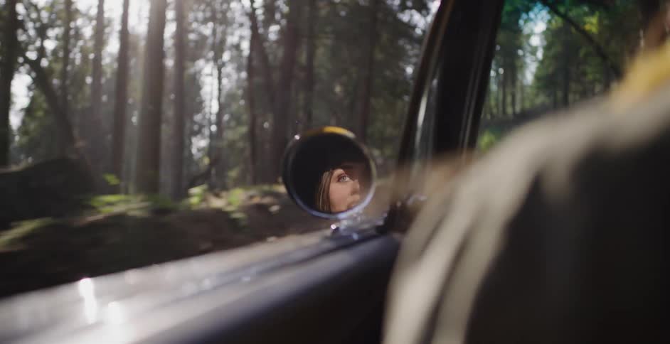A woman's expression of joy turns to fear in the reflection of a car's side view mirror as it travels down a winding forest road