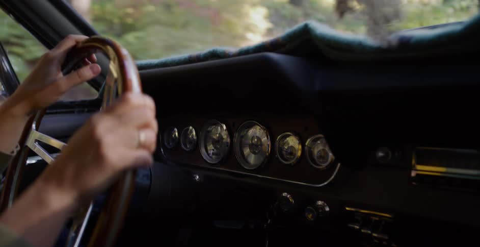 Close up of a driver's hands gripping the wooden steering wheel of a classic car as they travel through a sunlit forest