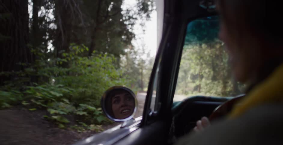 Over the shoulder view of an excited woman's reflection in a classic car's side-view mirror while driving through a forest