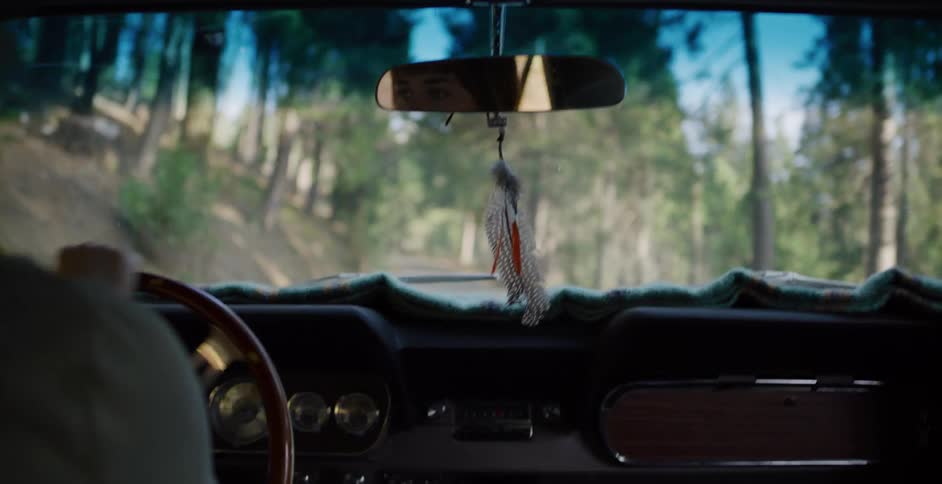 A woman's eyes reflect in the rearview mirror as she drives a vintage car down a bumpy forest road