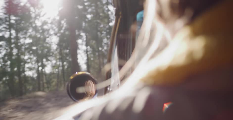 A woman's joyful reflection is seen in the side-view mirror of a vintage car driving through a sunlit forest
