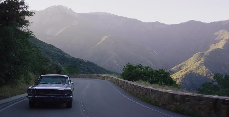 A classic black Mustang cruises down a winding mountain road in slow motion as the sun sets in the background