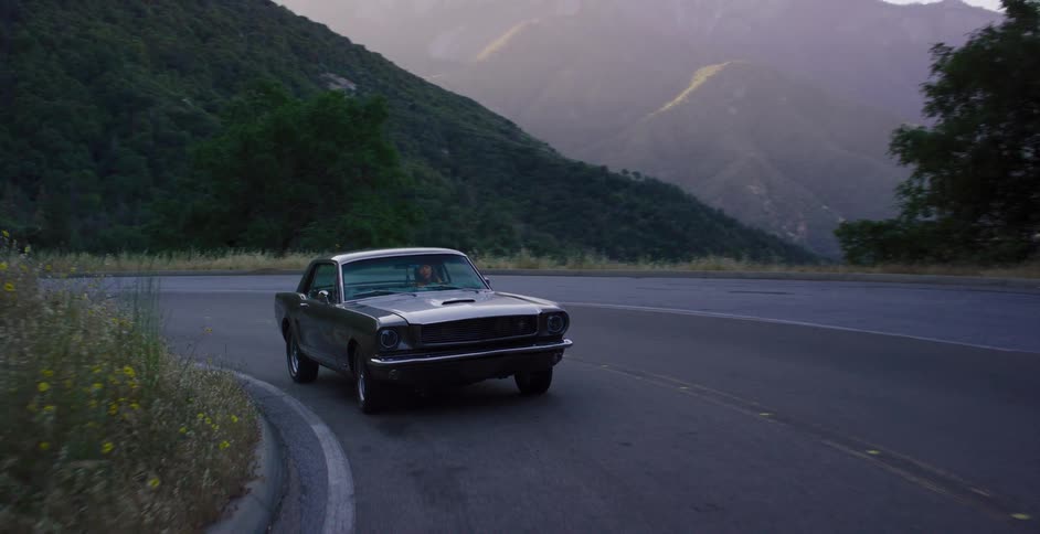 A vintage gray Ford Mustang cruises along a winding mountain road at dusk