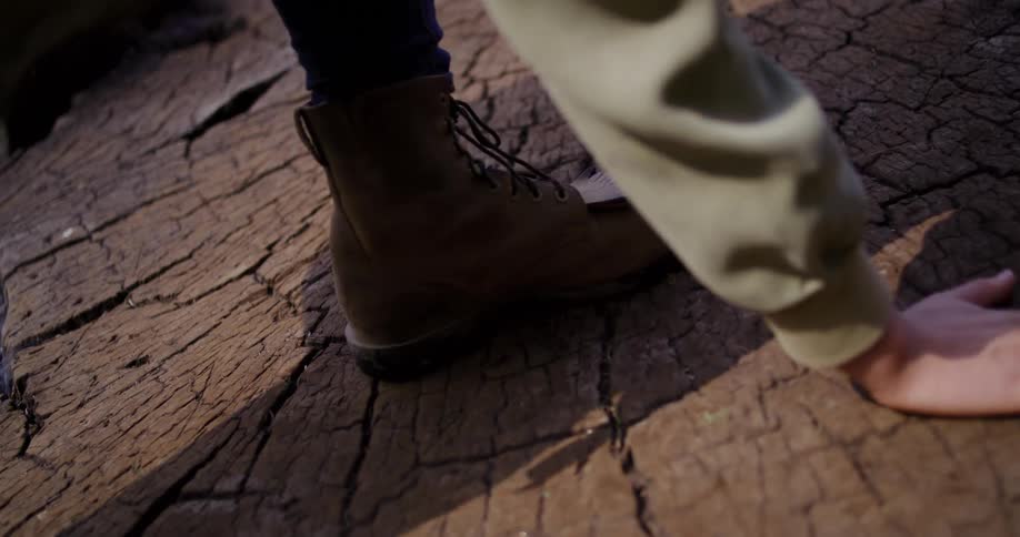 A person wearing hiking boots gently touches the textured top of a large tree stump before walking across it