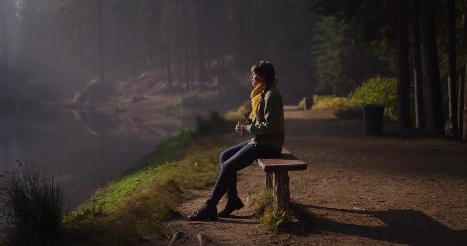 A woman relaxes on a wooden bench by a misty lake in the forest, drinking a warm beverage as the morning sun shines through the trees