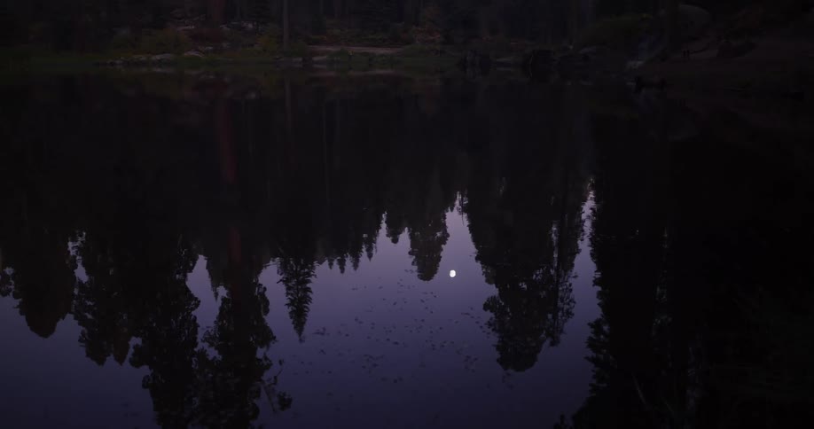 A slow pull out reveals the moon and a dark forest reflected on the serene surface of a lake at dusk