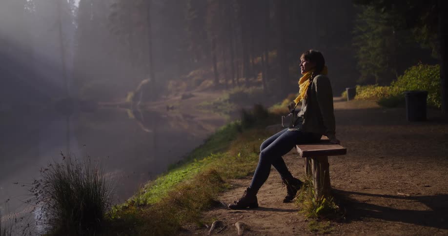 A young woman enjoys a moment of tranquility, sitting on a bench by a misty lake as beautiful sun rays shine through the forest