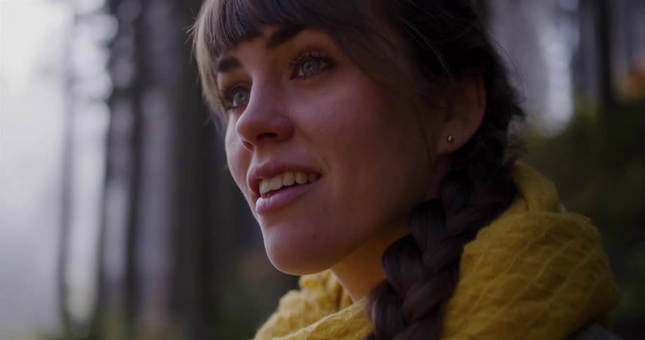 A beautiful young woman with a braid and a yellow scarf looks up with a happy smile while exploring a peaceful forest