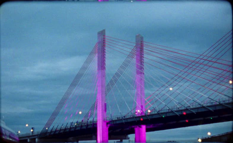 Vintage film captures a modern bridge at dusk as its towers glow with changing blue and purple lights, ending in an abstract blur