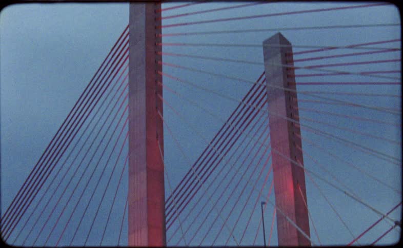 A low-angle shot tilts up the towers of a large suspension bridge illuminated with pink lights against the dusky sky
