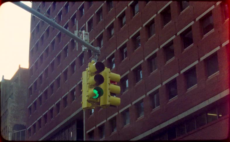 A retro, film-style shot of a city traffic light changing from red to green in front of a brick building, with a film burn transition