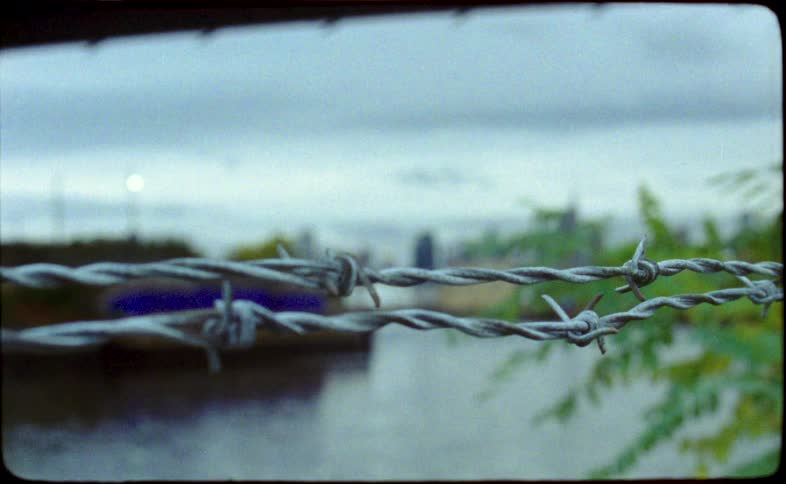 Vintage Super 8 film footage captures a close-up of barbed wire with an out-of-focus industrial canal in the background