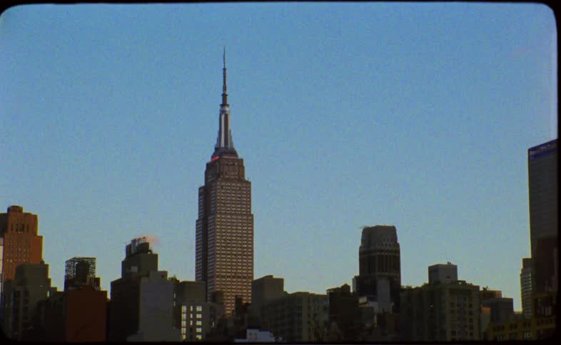 A vintage film shot captures the Empire State Building towering over the New York City skyline against a clear blue sky at sunset
