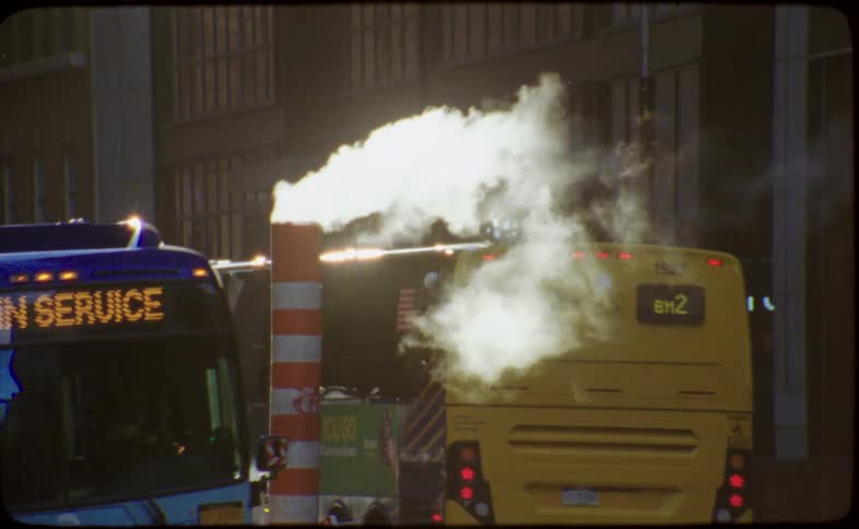 Steam rises from a vent on a busy New York City street as buses pass by, captured in a vintage film style