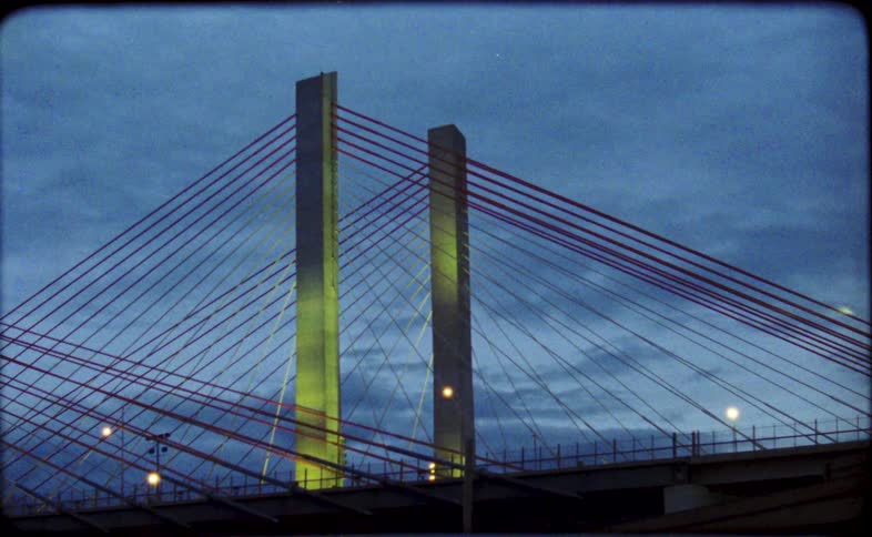 A retro, film-style shot of a cable-stayed bridge with its towers illuminated in changing colors at dusk, seen from a moving vehicle creating abstract light trails