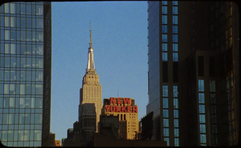 A vintage film shot captures the iconic Empire State Building and the New Yorker Hotel sign framed between modern skyscrapers in New York City