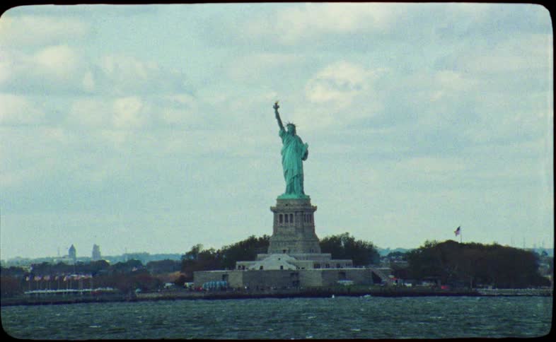A grainy, vintage film shot shows the Statue of Liberty from the water on a cloudy day in New York City