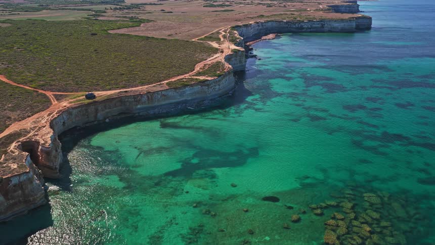 An epic aerial view pulls back to reveal a stunning, rugged coastline with sheer cliffs and a clear, turquoise sea on a sunny summer day
