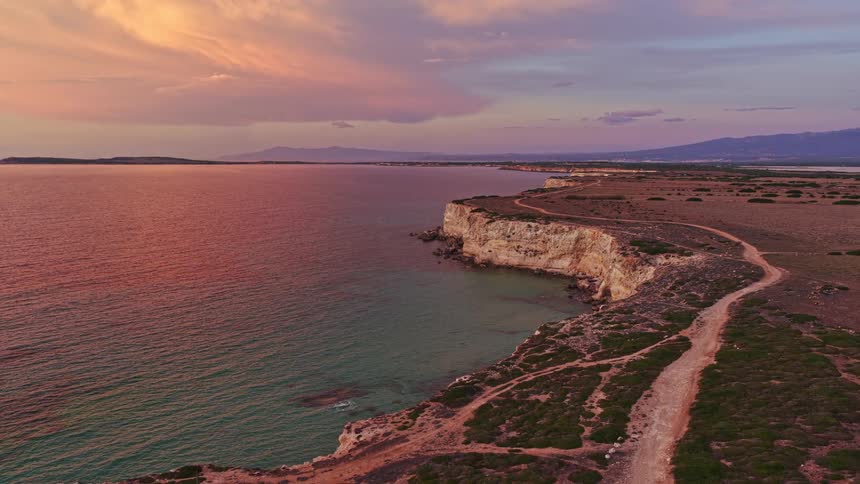 A breathtaking aerial view of a rugged coastline with dramatic cliffs and winding dirt paths under a magnificent pink and purple sunset sky
