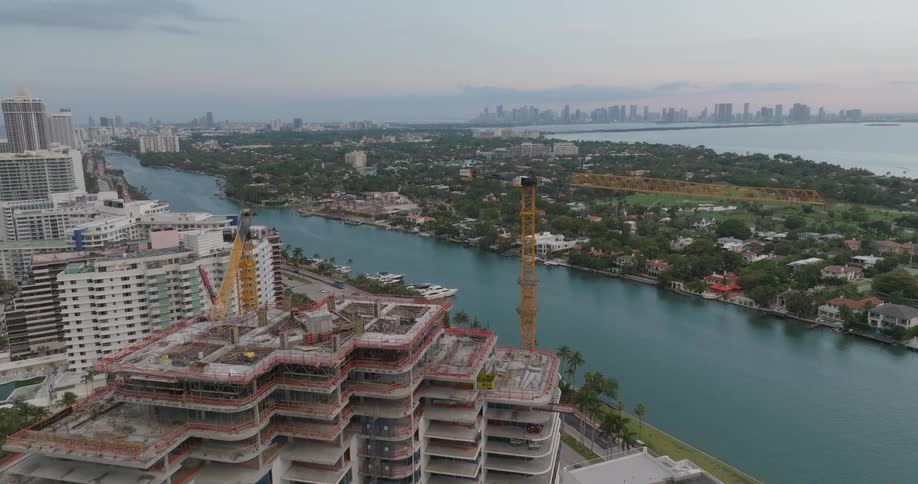 Aerial footage circles a high-rise construction site with large cranes on the Miami Beach waterfront, with the downtown skyline in the distance