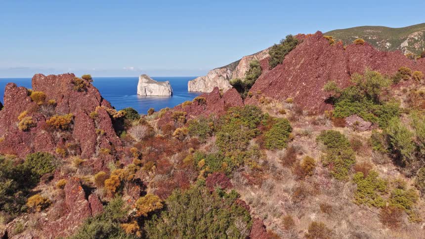 A dramatic aerial reveal of the Pan di Zucchero sea stack, flying over the rugged red cliffs of the Sardinian coastline