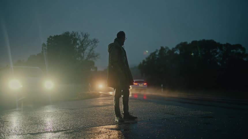 A lonely man stands in the rain on a dark road at night, illuminated by the headlights of a car as traffic passes by