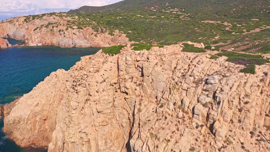 An aerial view pulls back from a secluded turquoise cove to reveal a stunning and rugged rocky coastline on a sunny day