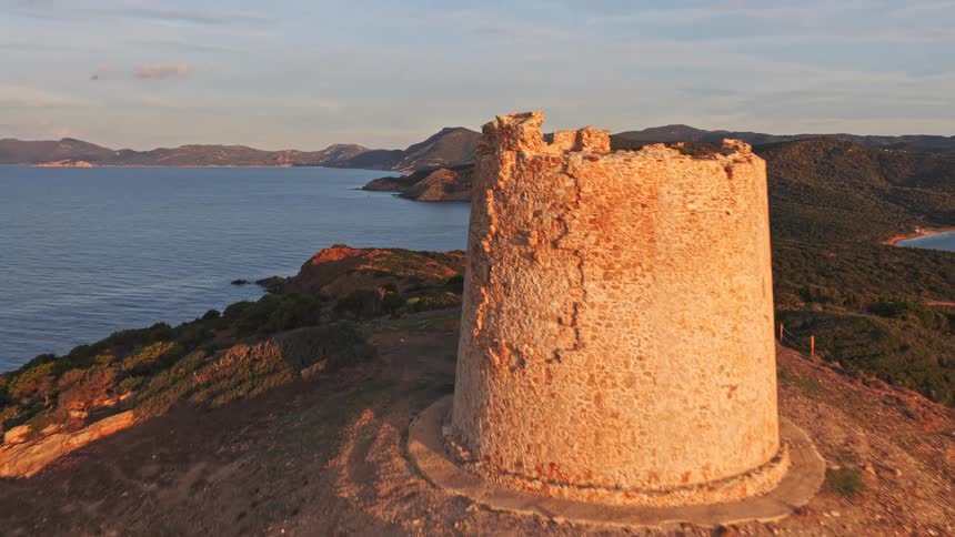 Stunning aerial footage flies past a ruined coastal watchtower on a hilltop, revealing the beautiful Sardinian coastline at sunset
