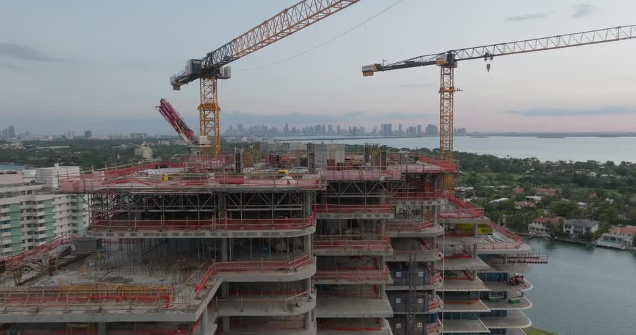 An aerial shot pulls back from a coastal high-rise building under construction with two large cranes at dusk