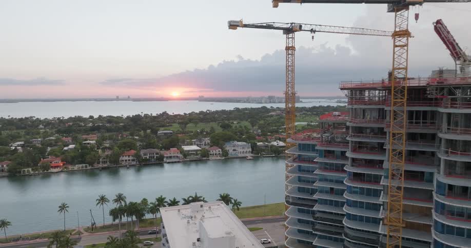 Aerial pullback revealing a waterfront skyscraper under construction with large cranes against a beautiful sunset backdrop