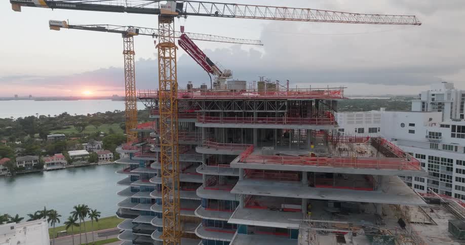 Slow aerial approach to a modern skyscraper under construction with large cranes overlooking the city and water at sunset