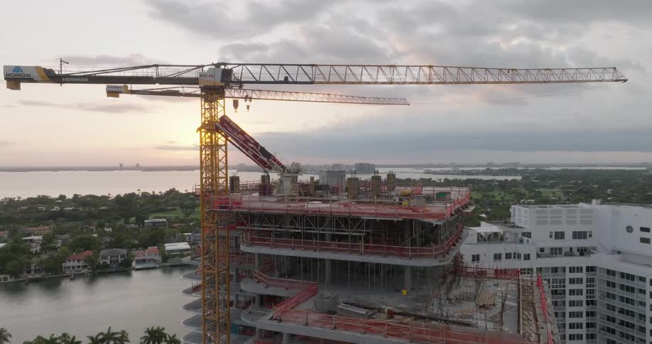 Drone ascends to reveal a large coastal construction project with towering cranes against a beautiful sunset sky