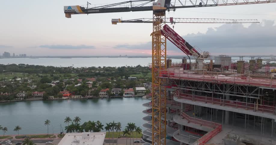 Aerial orbit of a skyscraper under construction with large cranes overlooking the water and city skyline at dawn