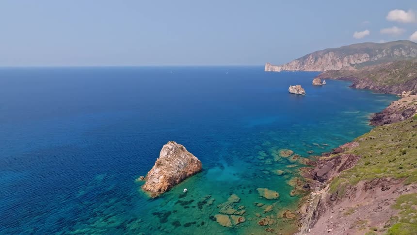 A beautiful aerial view pans across a rugged coastline, revealing stunning rock formations in the crystal-clear turquoise and deep blue sea
