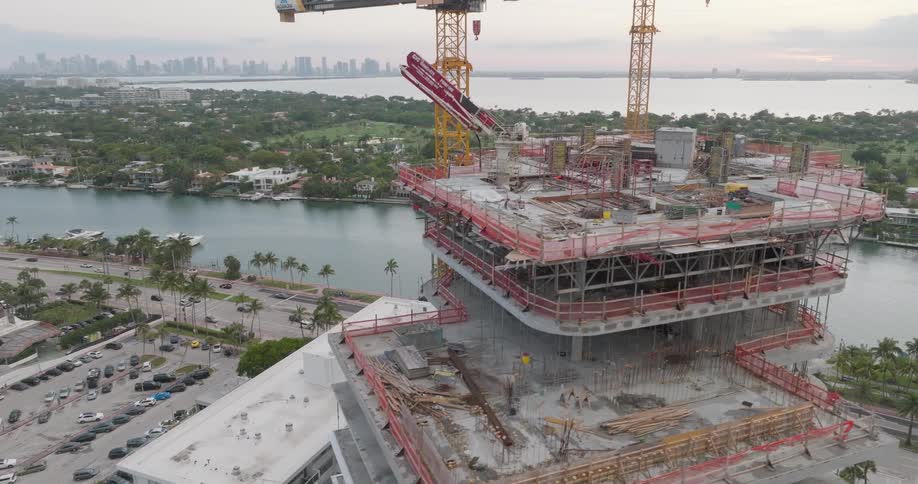 Aerial view reveals a modern high-rise building construction site with cranes overlooking the water and a city skyline