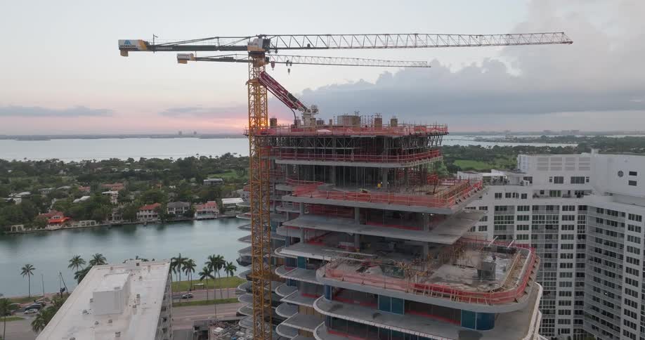 An ascending aerial shot reveals a modern skyscraper under construction with large cranes overlooking a coastal city at sunset