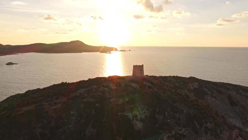Cinematic aerial pullback reveals a historic stone watchtower on a rugged island peninsula during a beautiful golden sunset over the sea