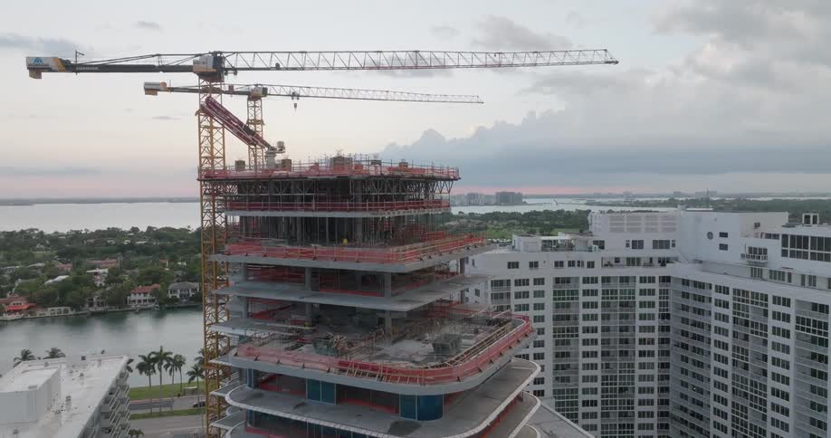 Aerial view ascending alongside a modern high-rise building under construction with tower cranes overlooking a coastal city at dusk