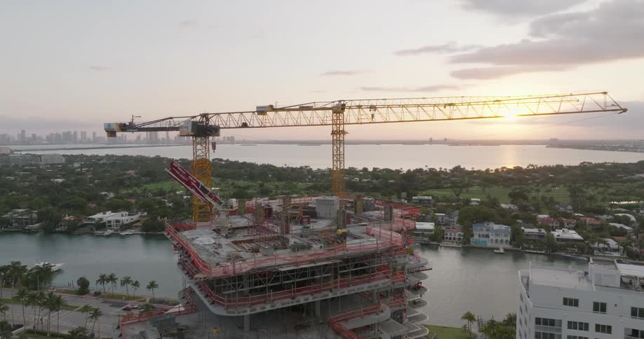 Aerial view moving towards a waterfront high-rise construction site with cranes during a golden sunset