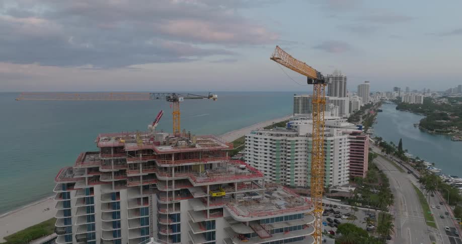 A slow aerial orbit reveals a luxury high-rise condominium under construction with cranes on the Miami Beach waterfront at dusk