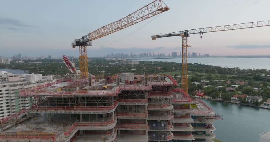 An aerial pullback shot reveals a large skyscraper being built with cranes overlooking a coastal city skyline at dusk
