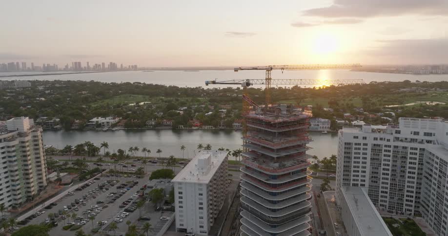 Aerial orbit of a modern high-rise building under construction with cranes against a beautiful sunset over the Miami skyline and waterway