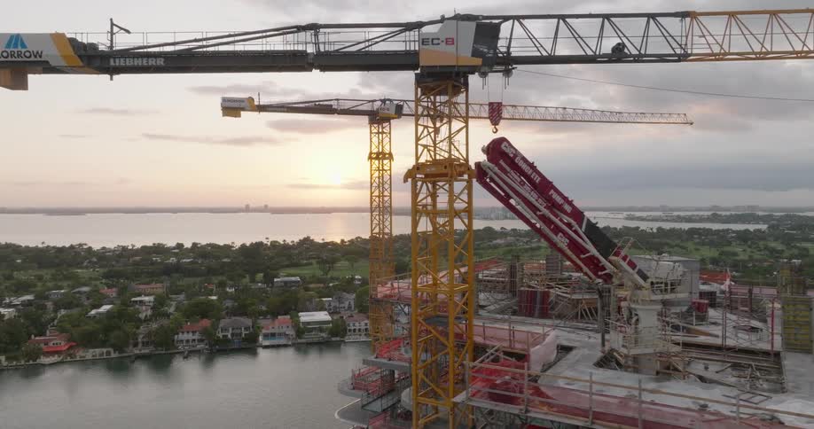 Towering construction cranes operate on a waterfront high-rise building site against a beautiful sunset backdrop