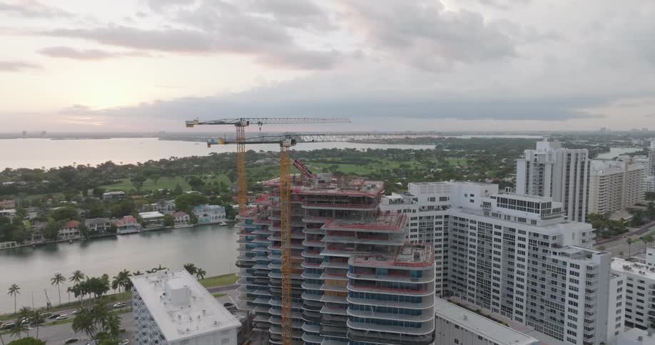 Aerial footage circles a towering skyscraper under construction on the Miami Beach coast at sunrise, revealing the cityscape and ocean