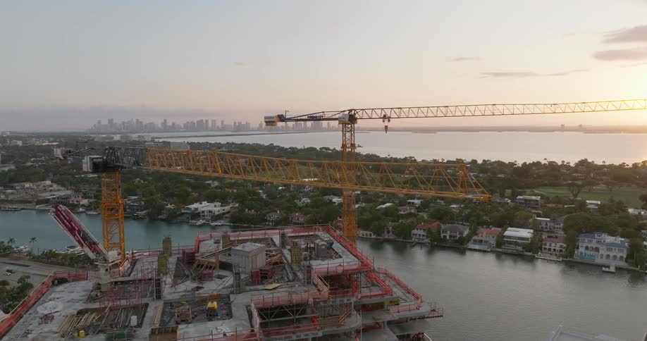Cinematic aerial footage flies towards a high-rise building under construction with large cranes overlooking the Miami, Florida skyline at sunrise